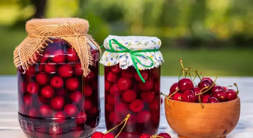 Dwa słoje domowego kompotu z czereśni, jeden z lnianym, drugi z kwiecistym nakryciem, obok miseczka świeżych owoców.