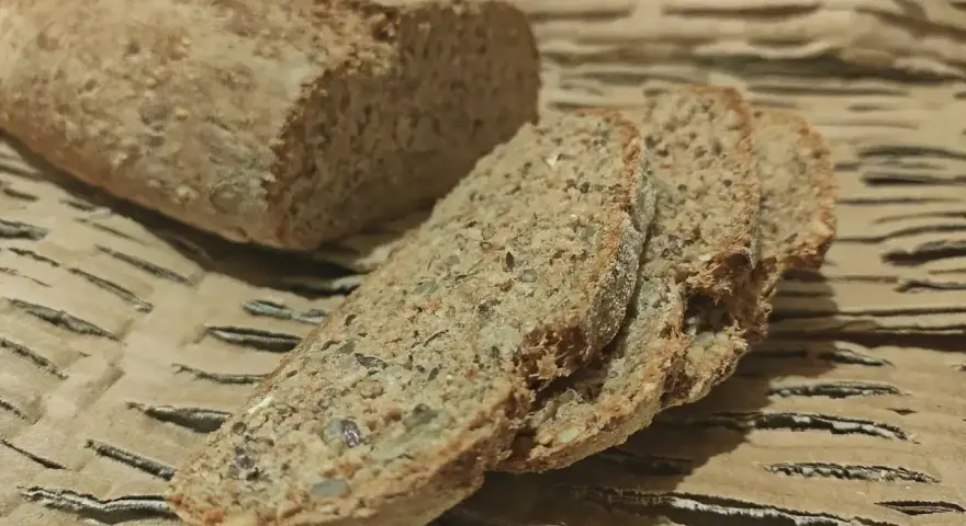 Chleb orkiszowy, pokrojony na kromki, z widocznymi ziarnami, leży na falistej tekturze.