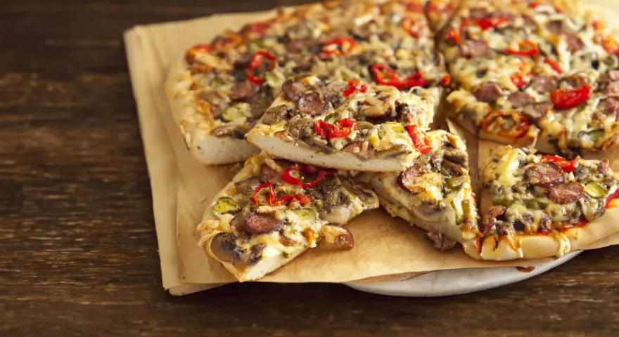 Grube ciasto na pizze z kiełbasą, pieczarkami i papryką, podane na papierze do pieczenia.