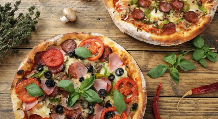 Dwie pizze z dodatkami: salami, pomidory, oliwki, szynka, pieczarki i bazylia.