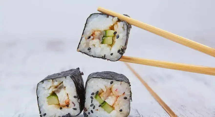 Trzy kawałki sushi z ryżem, ogórkiem i rybą, jeden podniesiony pałeczkami. Idealne do sosu, który stanowi zalewa do sushi.