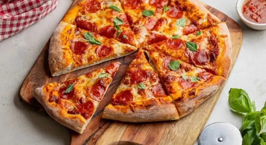 Pikantna pizza diavola z pepperoni i bazylią, pokrojona na kawałki, leży na drewnianej desce. Obok sos i nóż do pizzy.