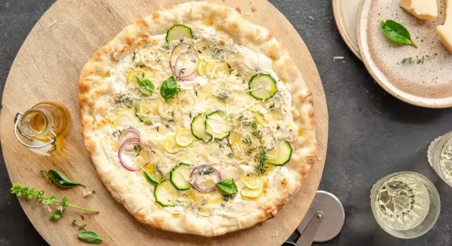 Pyszna pizza bianca z cukinią, cebulą i ziołami, podana na drewnianej desce. Obok buteleczka oliwy i kieliszki z winem.