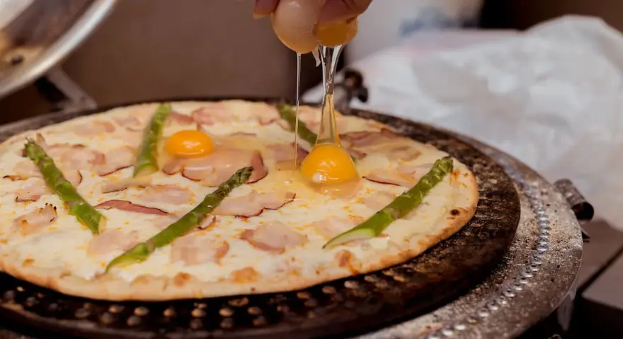 Ktoś dodaje jajko do pizzy z boczkiem i szparagami. Pyszna pizza z jajkiem w przygotowaniu.