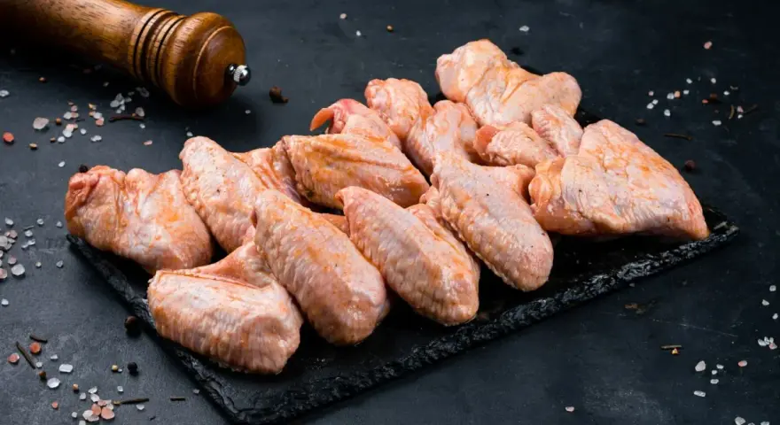 Skrzydełka kurczaka gotowe do pieczenia, zamarynowane w aromatycznej solance. Obok młynek do pieprzu i gruboziarnista sól.