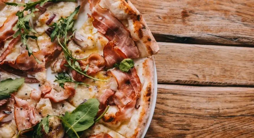 Pyszna pizza carbonara z chrupiącym boczkiem i rukolą, podana na drewnianym blacie.