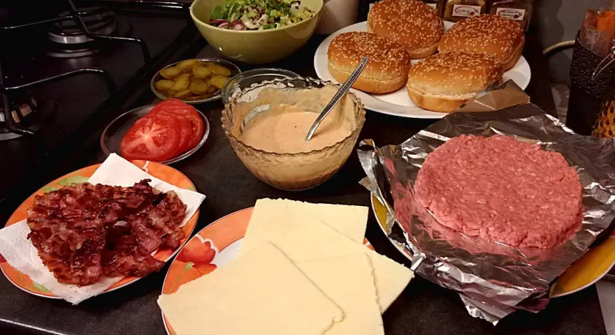 Składniki do przygotowania idealnego cheeseburgera: bułki, surowe mięso, ser, bekon, pomidory, ogórki i sos.