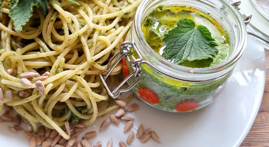 Makaron z pesto z pokrzywy, ozdobiony świeżymi liśćmi pokrzywy i pestkami słonecznika. Obok słoiczek z domowym pesto.