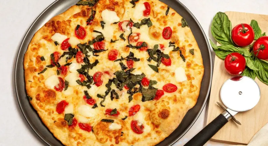 Pyszna pizza z serka wiejskiego, pomidorkami koktajlowymi i bazylią. Obok leży krajalnica do pizzy i świeże pomidory.
