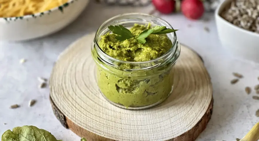 Pesto z liści rzodkiewki w słoiczku, ozdobione natką pietruszki, na drewnianym krążku. Obok czosnek, rzodkiewki i nasiona.
