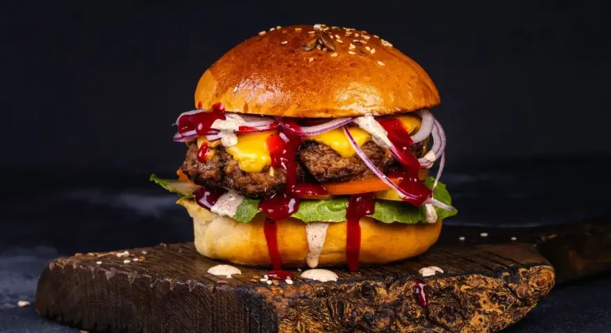 Soczyste burgery z grilla, polane sosem, z serem, cebulą i sałatą, na drewnianej desce.