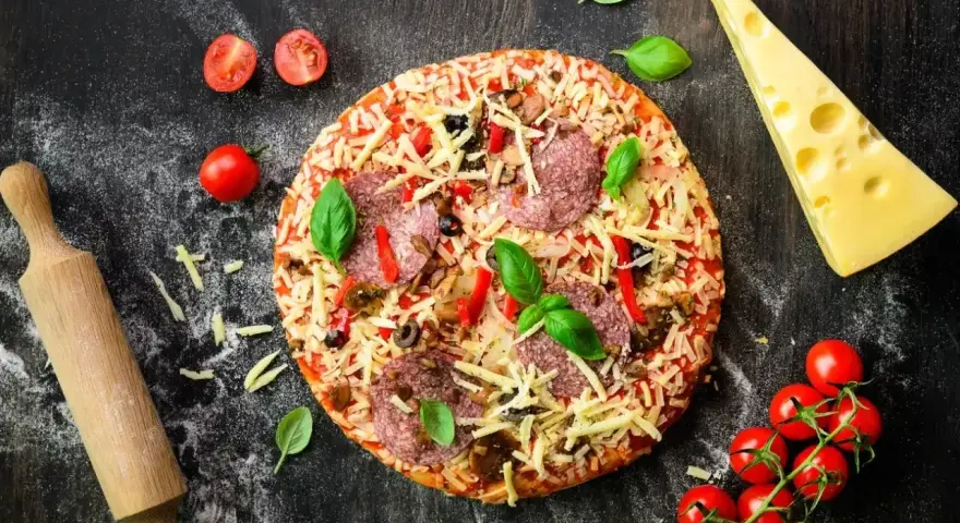 Gotowa pizza z salami, serem i warzywami, obok wałek, pomidorki i ser. Idealna na wieczór z przyjaciółmi, gdy brakuje czasu na przygotowanie ciasta z mąki na pizzę.