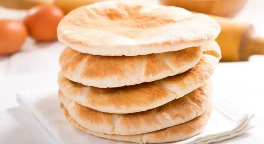 Stos świeżo upieczonego chleba arabskiego na białej serwetce, obok jajka i wałka do ciasta.