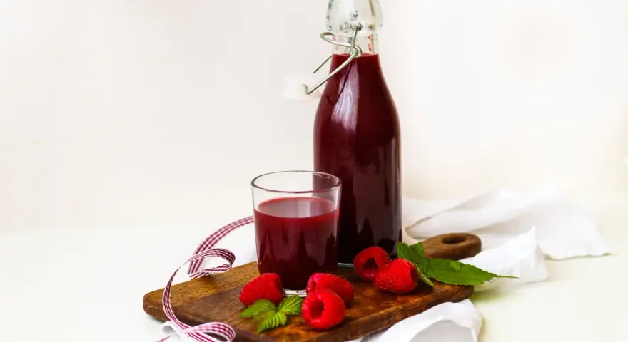 Szklanka i butelka z ciemnoczerwonym sokiem z malin, na drewnianej desce z malinami i listkami mięty.