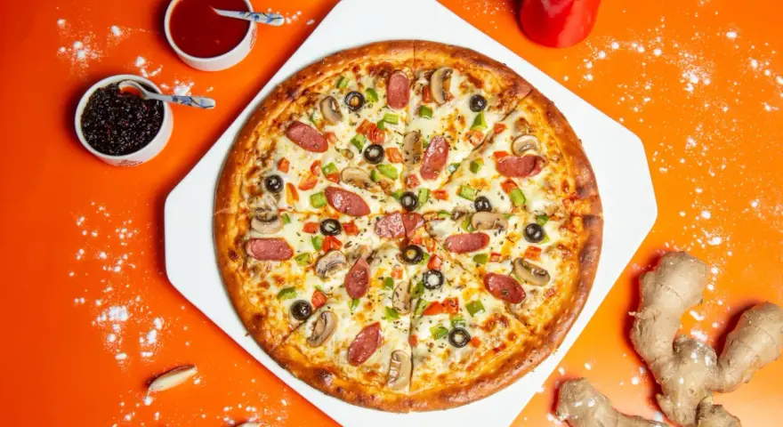 Pizza z kiełbasą, pieczarkami i oliwkami na pomarańczowym tle, obok sosy i imbir.