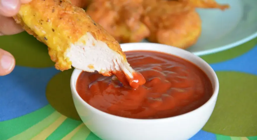 Chrupiący nugget zanurzany w gęstym, czerwonym sosie do nuggetsów. Na talerzu w tle widać więcej złotych kawałków kurczaka.
