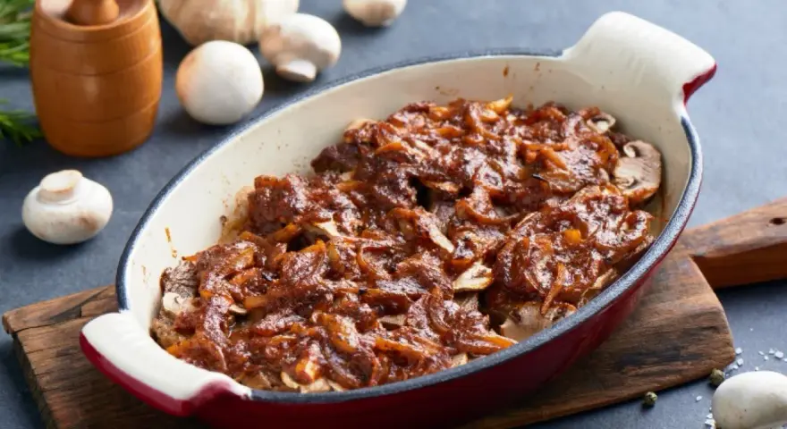 Karkówka pieczona w plastrach z cebulą i sosem, przypominającym ketchup. Danie w naczyniu żaroodpornym, obok grzyby i czosnek.