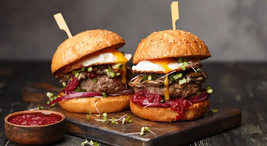 Dwa soczyste burgery z jajkiem sadzonym i grzybami. Idealny stopień wysmażenia burgera podkreśla smak. Obok miseczka sosu.