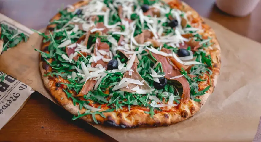 Apetyczna pizza prosciutto z rukolą, oliwkami i parmezanem, podana na papierze.