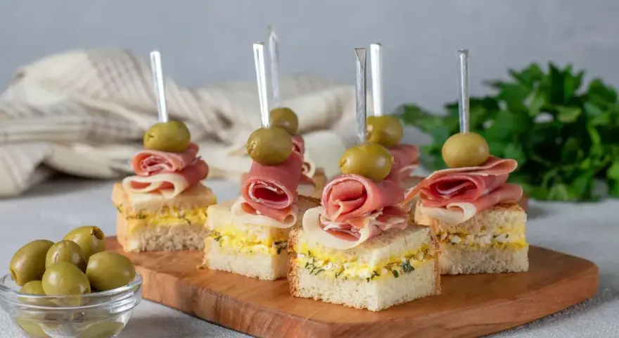 Mini kanapki z szynką, jajkiem i oliwkami na drewnianej desce. Idealne finger food na przyjęcie.