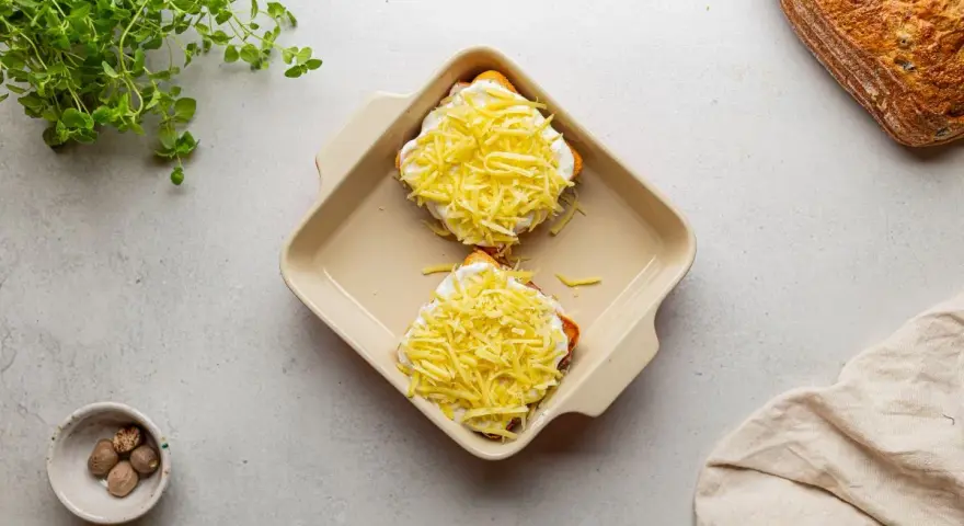 Dwa tosty z serem i beszamelem, przygotowane do zapieczenia jako croque madame. Obok gałka muszkatołowa i świeże zioła.