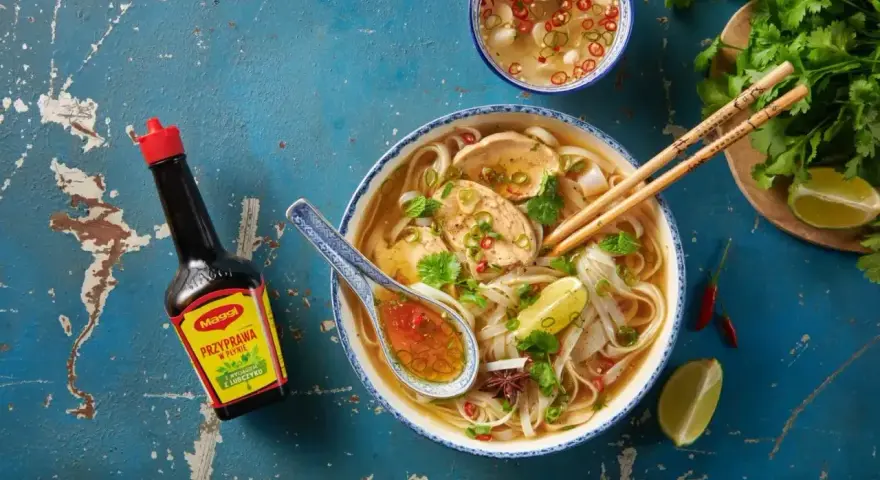 Aromatyczny makaron pho z kurczakiem, świeżą kolendrą, limonką i papryczkami chili. Obok butelka sosu Maggi.