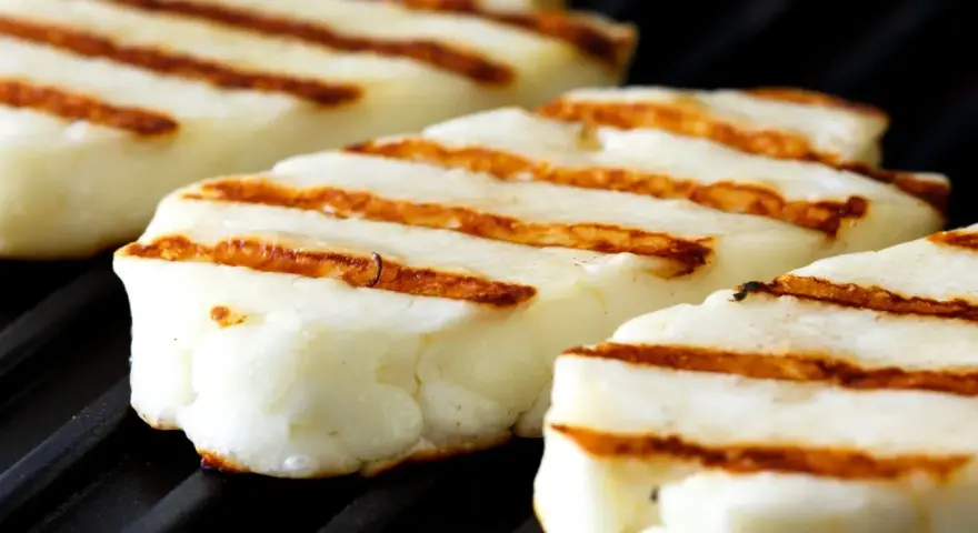 Trzy kawałki halloumi z grilla, z charakterystycznymi brązowymi paskami, leżą na czarnej, grillowej powierzchni.