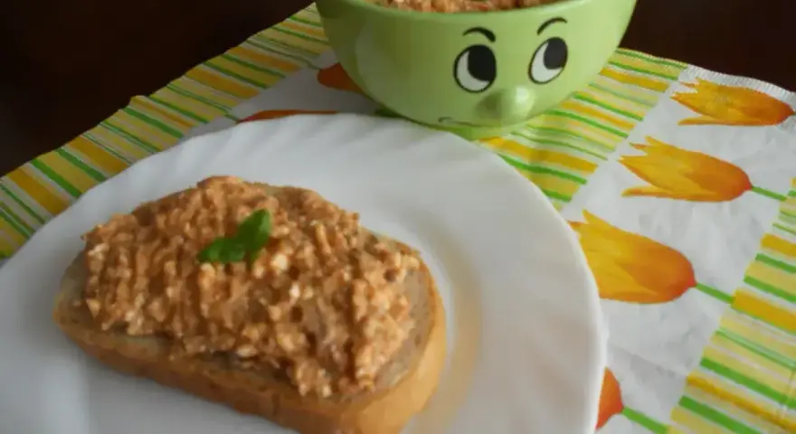 Pyszna pasta z makreli i twarogu na kromce chleba, udekorowana listkiem bazylii. Obok miseczka z pastą i wesołą buźką.