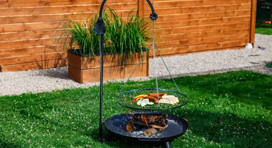 Grill trójnóg z paleniskiem i rusztem, na którym znajdują się kiełbaski, pieczywo i warzywa.