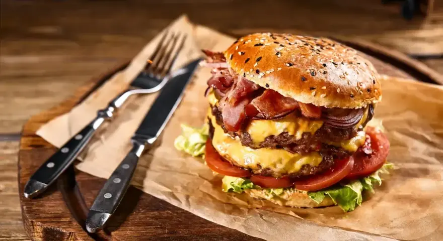 Podwójny burger z soczystym mięsem na burgery, serem, bekonem i warzywami, gotowy do zjedzenia z widelcem i nożem.