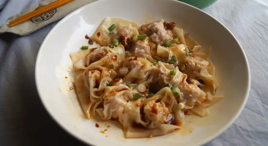 Pyszne ciasto wonton polane pikantnym sosem, posypane szczypiorkiem i sezamem. Obok pałeczki i miseczka z sosem.