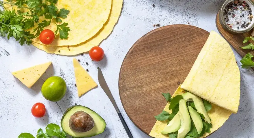 Keto tortilla z awokado i rukolą na drewnianej desce, obok pomidorki, ser i limonka.