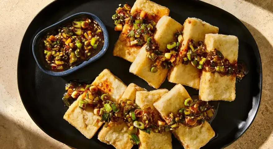 Chrupiące kawałki tofu polane aromatyczną marynatą z dymką i sezamem, podane na czarnym talerzu.