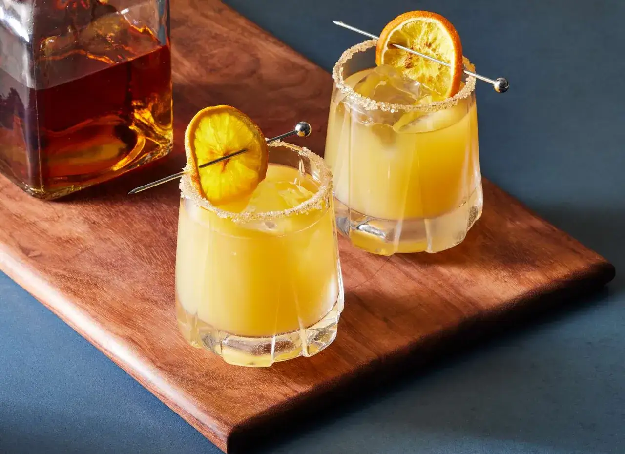 Dwa orzeźwiające drinki z pomarańczą i cukrowym rantem, idealne do przygotowania według przepisu na whisky sour. Obok butelka bursztynowego alkoholu.