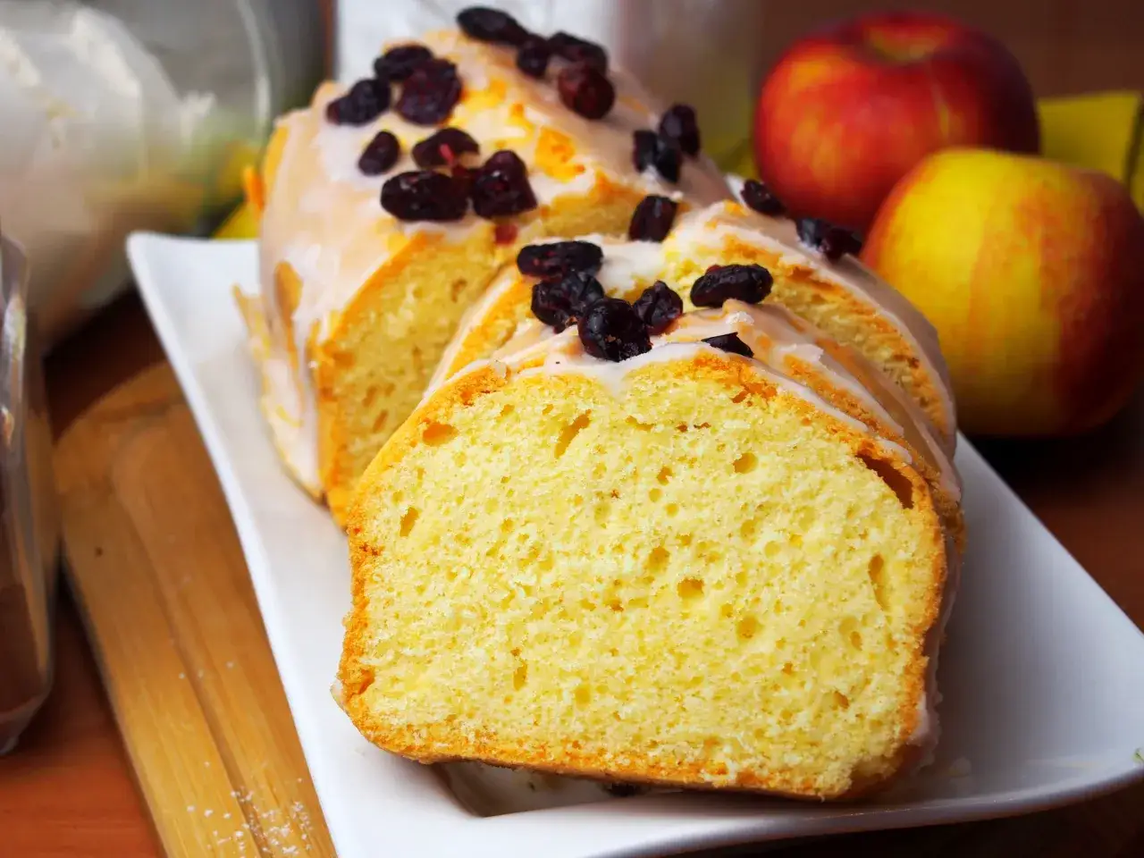 Wilgotna babka w keksówce, polana lukrem i posypana żurawiną, prezentuje się apetycznie obok jabłek.