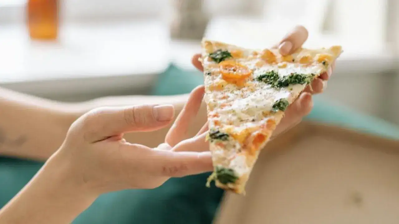 Dłonie trzymają kawałek pizzy z pomidorkami i szpinakiem. Idealny sposób, jak jeść pizzę, by delektować się każdym kęsem.