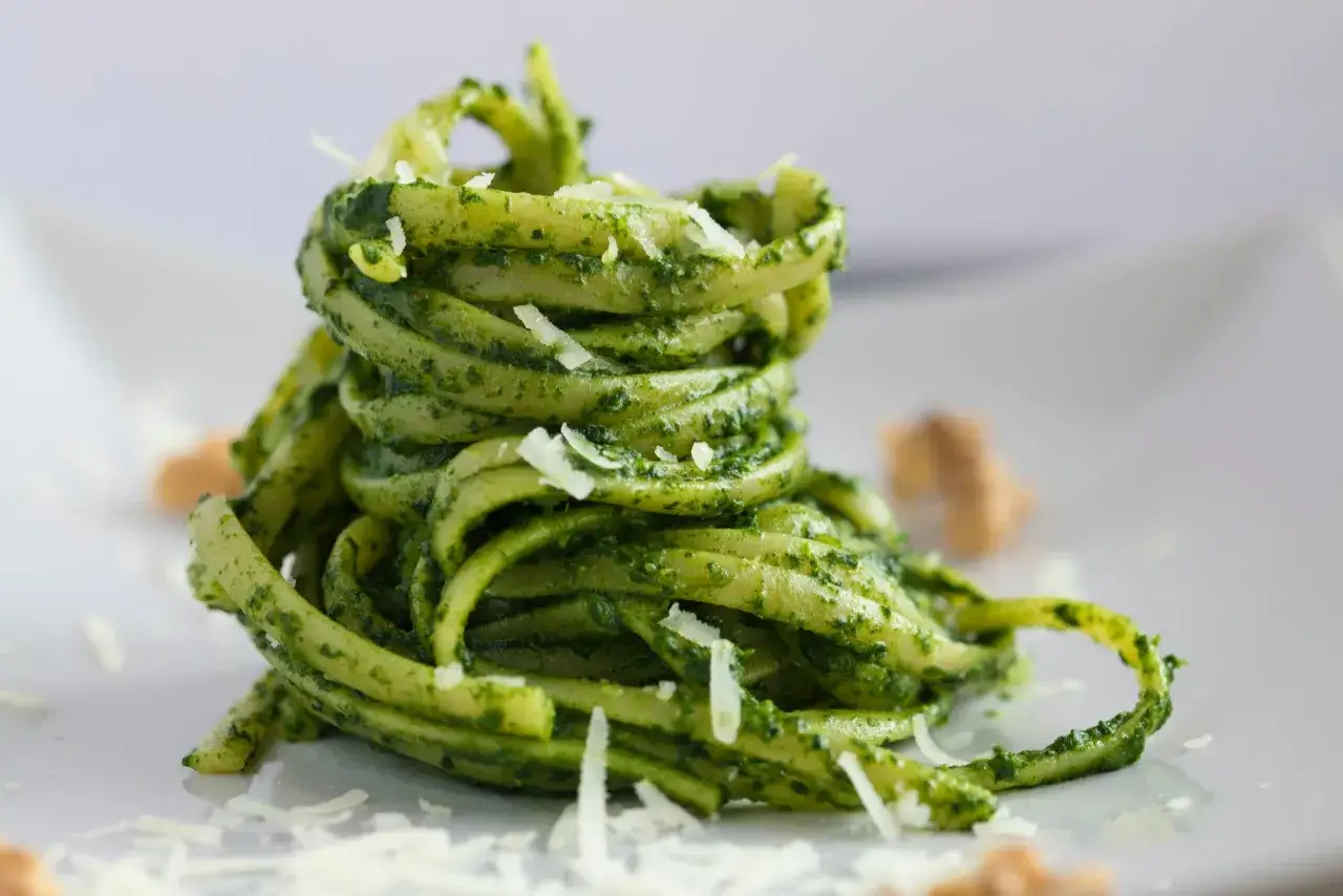 Stos włoskich dań z makaronem, polany zielonym pesto i posypany parmezanem.