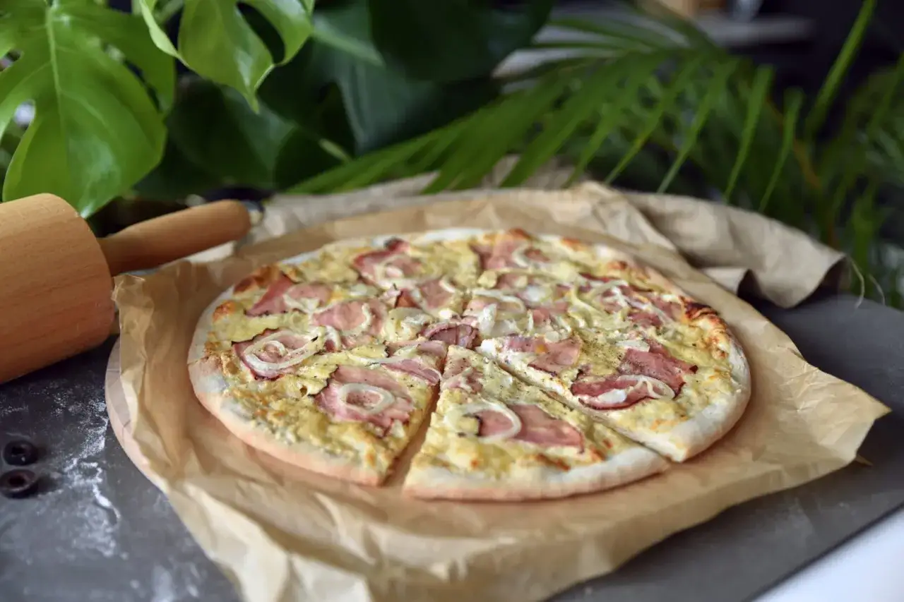 Pizza z szynką i cebulą, gotowa do podania. Widać składniki na wierzch: plastry szynki, krążki cebuli i ser. Obok wałek do ciasta.