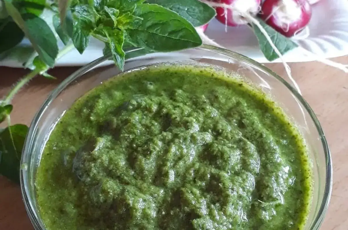 Pesto z liści rzodkiewki w szklanej miseczce, obok świeże liście i pęczek rzodkiewek.