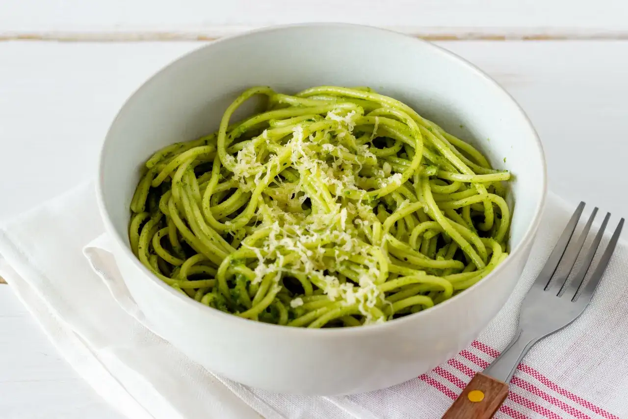 Miseczka spaghetti z intensywnym, zielonym pesto z pokrzywy, posypana startym serem. Obok leży widelec.