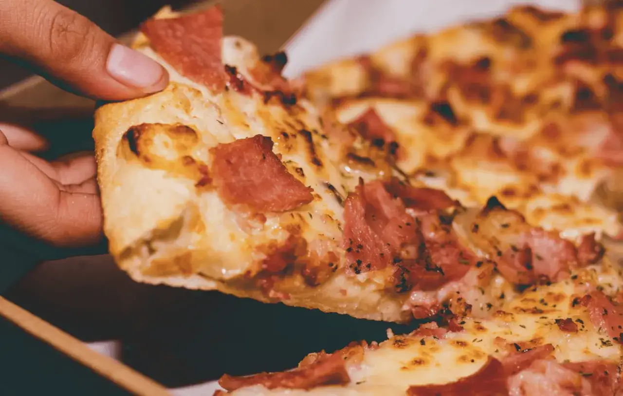 Kawałek pizzy z szynką i serem, pachnący sos umami, podnoszony palcami.