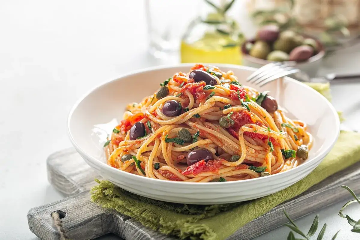 Pyszny makaron spaghetti z sosem pomidorowym, oliwkami i kaparami, podany w białej misce na zielonej serwetce.