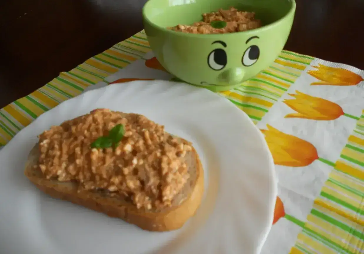Pyszna pasta z makreli i twarogu na kromce chleba, ozdobiona listkiem bazylii. Obok miseczka z pastą i zabawna buźka.