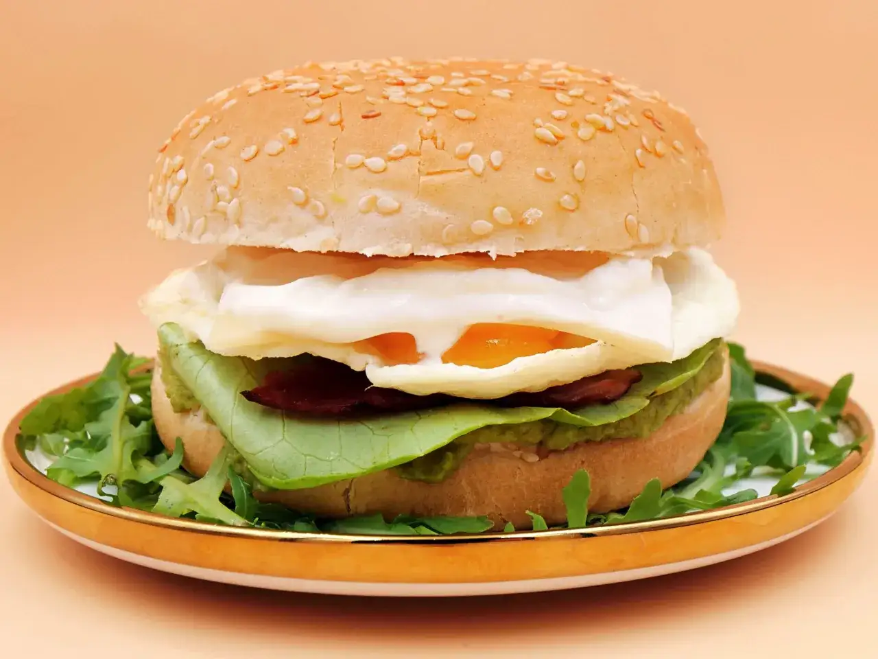 Burger z jajkiem sadzonym, bekonem i awokado. Pyszne dodatki do burgera na talerzu z rukolą.