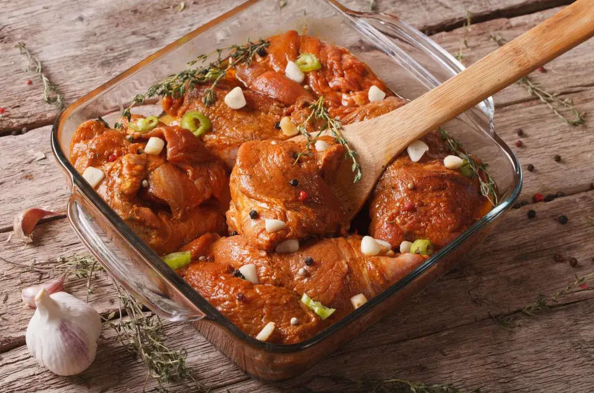 Mięso w szklanym naczyniu, zanurzone w aromatycznej marynacie z czosnkiem, ziołami i pieprzem. Drewniana łopatka miesza składniki.