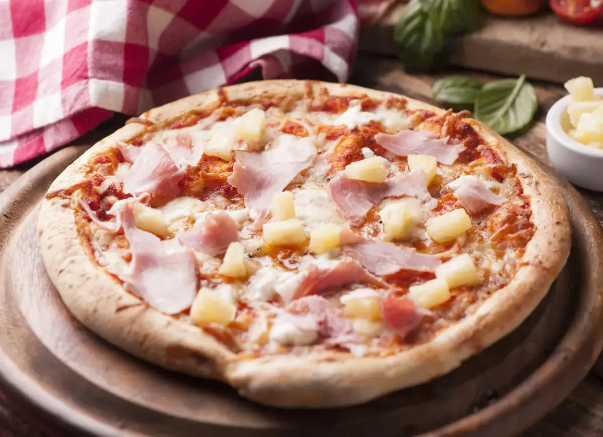 Hawajska pizza z szynką i ananasem, jedna z wielu pysznych pizz, prezentuje się apetycznie na drewnianej desce.