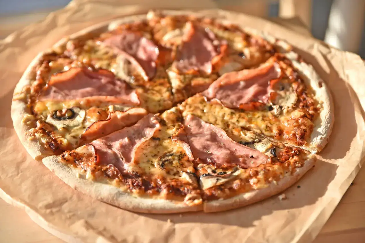 Pizza z szynką i pieczarkami, idealna na szybki obiad. Jakie pieczarki do pizzy? Te ciemne, pokrojone w plasterki, świetnie komponują się z serem i wędliną.
