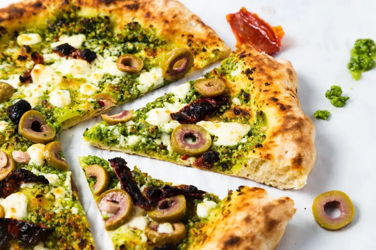 Kawałki pizzy z zielonym pesto, oliwkami i serem. Obok leżą suszone pomidory i dodatkowa porcja zielonego pesto.