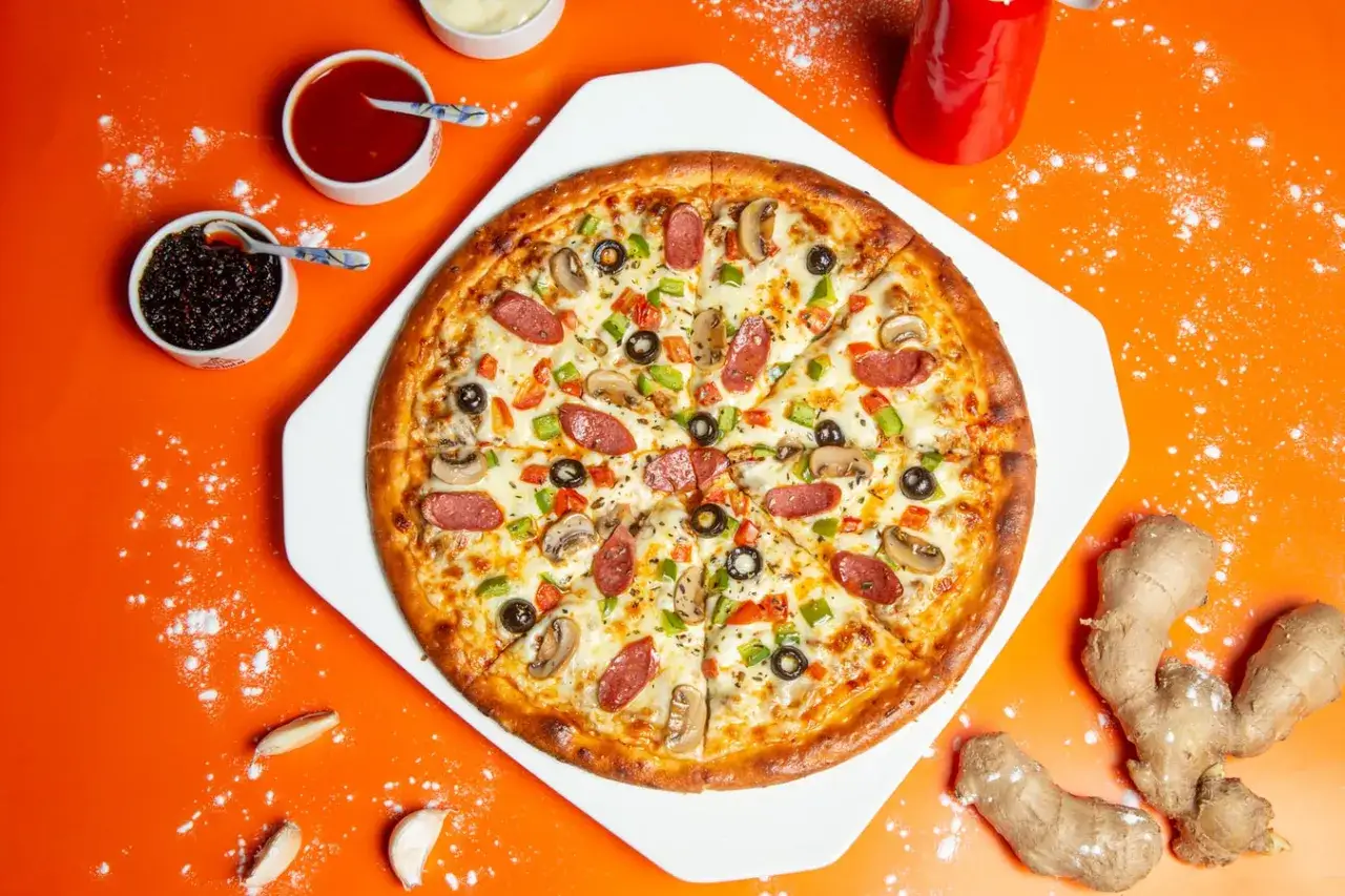 Pyszna pizza z pieczarkami, oliwkami i papryką. Na wierzchu widać plastry wędzonej kiełbasy, która świetnie komponuje się z resztą składników. To idealna pizza, jeśli zastanawiasz się, jaka szynka na pizzę będzie najlepsza!
