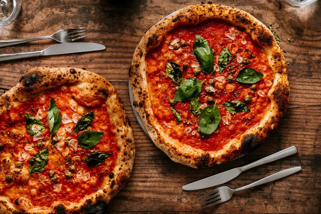 Dwie pizze neapolitańskie z pomidorami, bazylią i czosnkiem. Niezbędny dodatek smakowy włoskiej pizzy, gotowe do podania z widelcem i nożem.